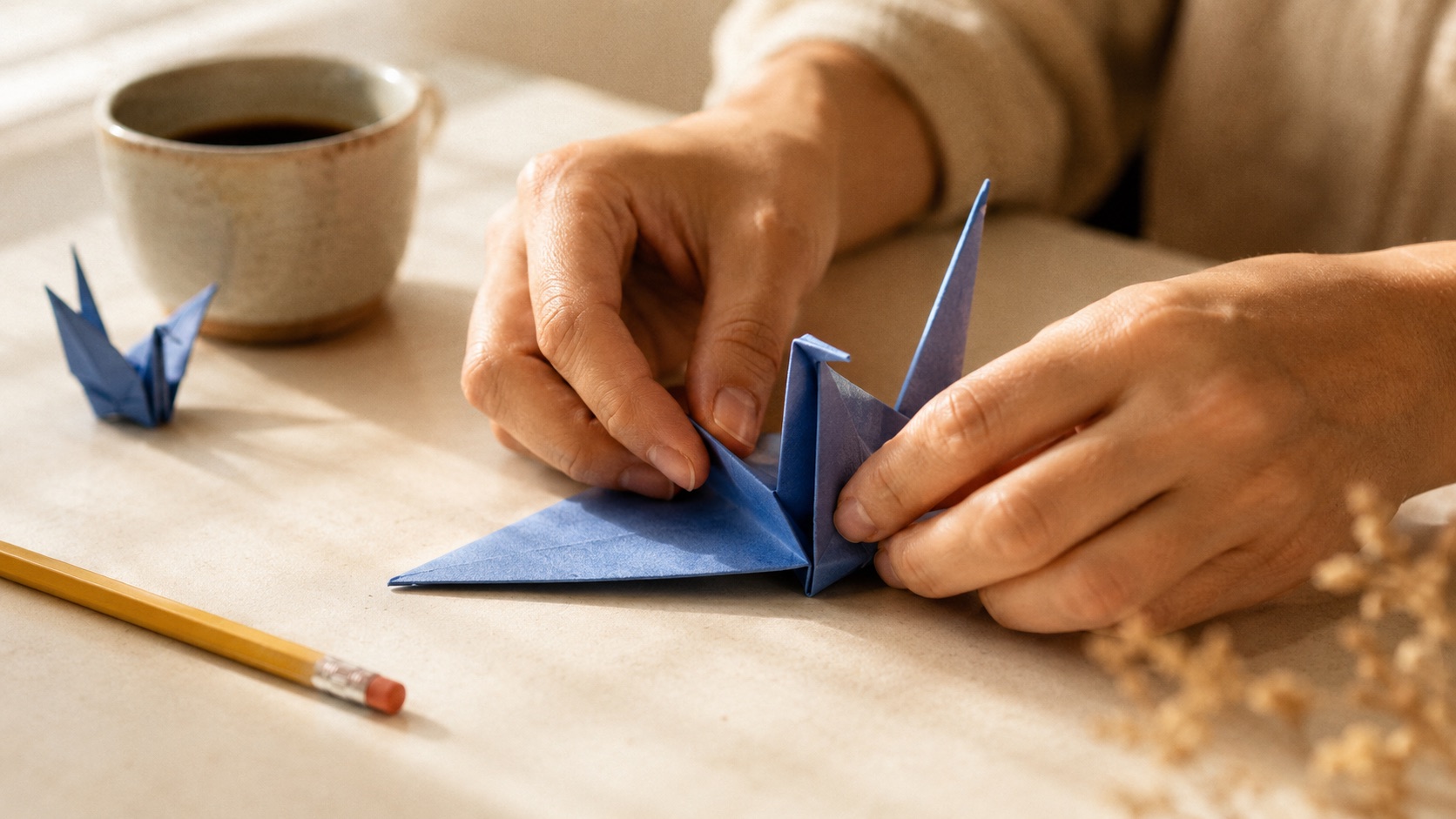 A pair of hands folds a periwinkle paper origami crane at a warm desk, a finished crane and a cup of coffee beside them.